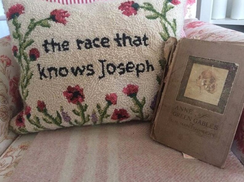 K&ouml;nnte beinhalten: Ein dekoratives Kissen mit dem Text "the race that knows Joseph", umgeben von floralen Stickereien. Daneben liegt ein Vintage-Buch mit dem Titel "Anne of Green Gables" von L.M. Montgomery mit verblichenem Einband.