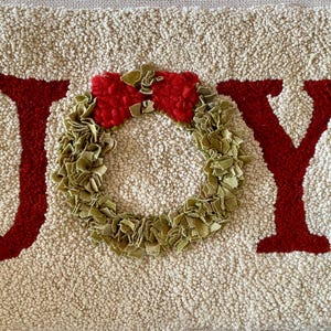 May include: A cream-coloured rug with the word "JOY" in red, featuring a green wreath with a red bow. The rug has a textured, looped pile, creating a cosy, festive feel. Ideal for holiday decor.