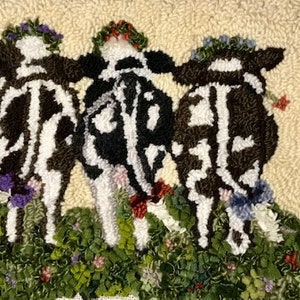 May include: A handmade rug depicting three cows with black and white patterns, each wearing floral wreaths. The cows are positioned in a field of green and colorful flowers, set against a cream backdrop.