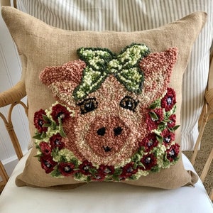 May include: A decorative burlap pillow featuring a hand-stitched pig face design. The pig has a green bow, a floral wreath, and black eyes. The pillow is placed on a white chair with a striped background.