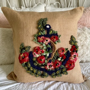 May include: Square beige burlap pillow with a floral design. The design features red flowers, green leaves, and a dark blue border. The pillow is placed on a white bed.