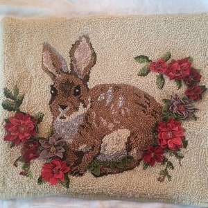 May include: A square, beige, textured pillow with a detailed, embroidered design. The design features a brown rabbit surrounded by red, purple, and green floral accents. The rabbit has long ears and is centered on the pillow.