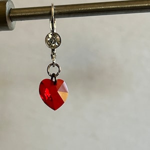 Pode incluir: Um brinco prateado com fecho de alavanca, apresentando um pequeno cristal transparente de corte redondo sobre um pendente de cristal vermelho vibrante em forma de coração. O brinco está suspenso numa fina barra metálica horizontal.