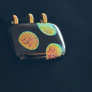 May include: A square pendant with a black base and three gold-colored loops at the top. The pendant features three iridescent, circular designs in shades of orange, yellow, and green. The pendant is displayed against a black background.