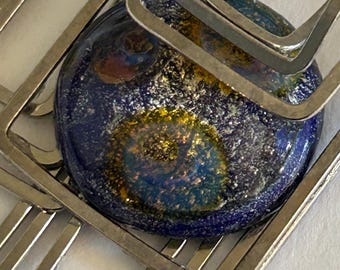 May include: Close-up of a decorative glass bead with a deep blue background and flecks of gold and silver. The bead is partially framed by a silver metal geometric design, creating a modern, abstract aesthetic. The bead's design resembles a galaxy.