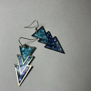 Puede incluir: Un par de pendientes con un diseño geométrico en capas. Cada pendiente presenta tres triángulos azules y turquesas con bordes de metal plateado. Los pendientes tienen un cierre de gancho.
