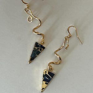 Può includere: Un paio di orecchini pendenti color oro con un design a filo metallico ondulato e serpentino collegato a un ciondolo triangolare. I ciondoli sono blu scuro con venature dorate irregolari, sospesi a ganci per orecchini standard.