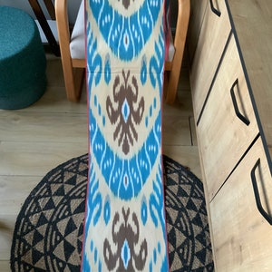 May include: A long piece of fabric with a blue, white, and brown ikat pattern. The fabric has a red border and is folded in half.