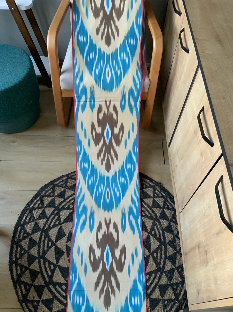 May include: A long piece of fabric with a blue, white, and brown ikat pattern. The fabric has a red border and is folded in half.