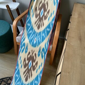 May include: A long, narrow fabric with a blue and white ikat pattern. The fabric has a red border and is draped over a wooden chair.