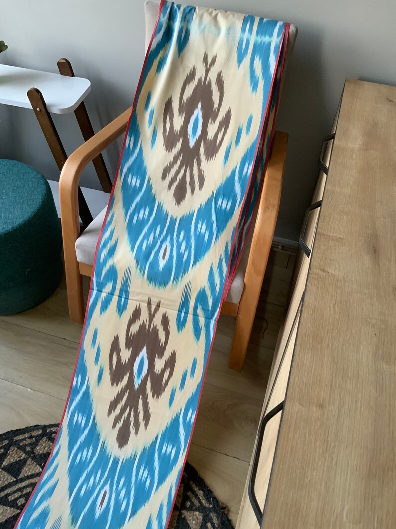 May include: A long, narrow fabric with a blue and white ikat pattern. The fabric has a red border and is draped over a wooden chair.