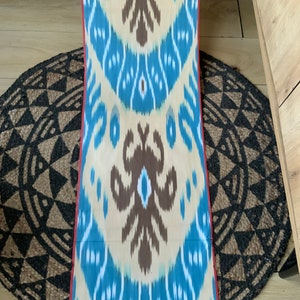 May include: A long, narrow piece of fabric with a repeating pattern of blue, white, and brown. The fabric has a red border on the edges.