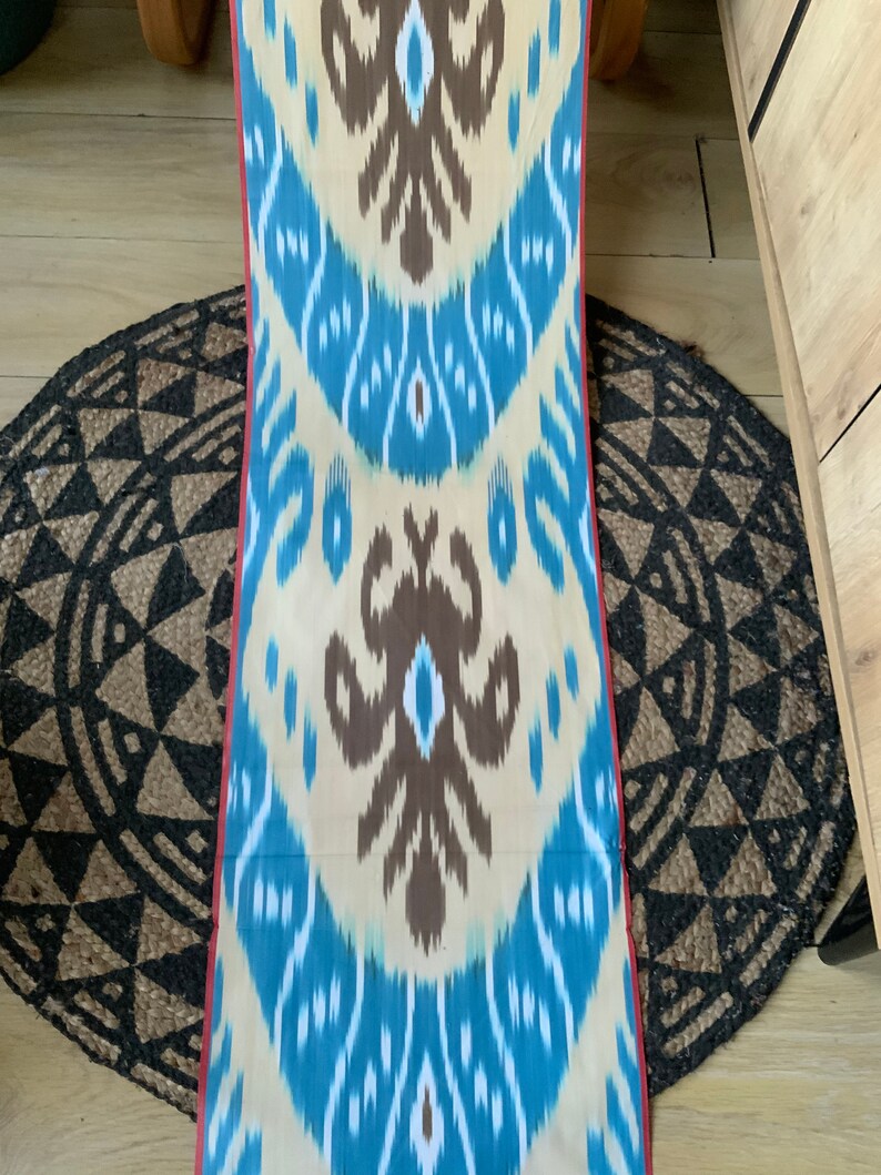 May include: A long, narrow piece of fabric with a repeating pattern of blue, white, and brown. The fabric has a red border on the edges.