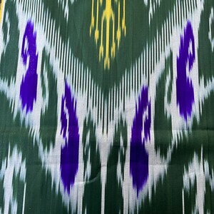 May include: Close-up of a fabric with a repeating ikat pattern. The design features a dark green background with vertical white and yellow lines. Bold purple shapes are interspersed throughout the pattern, creating a striking visual effect. The fabric has a textured appearance.
