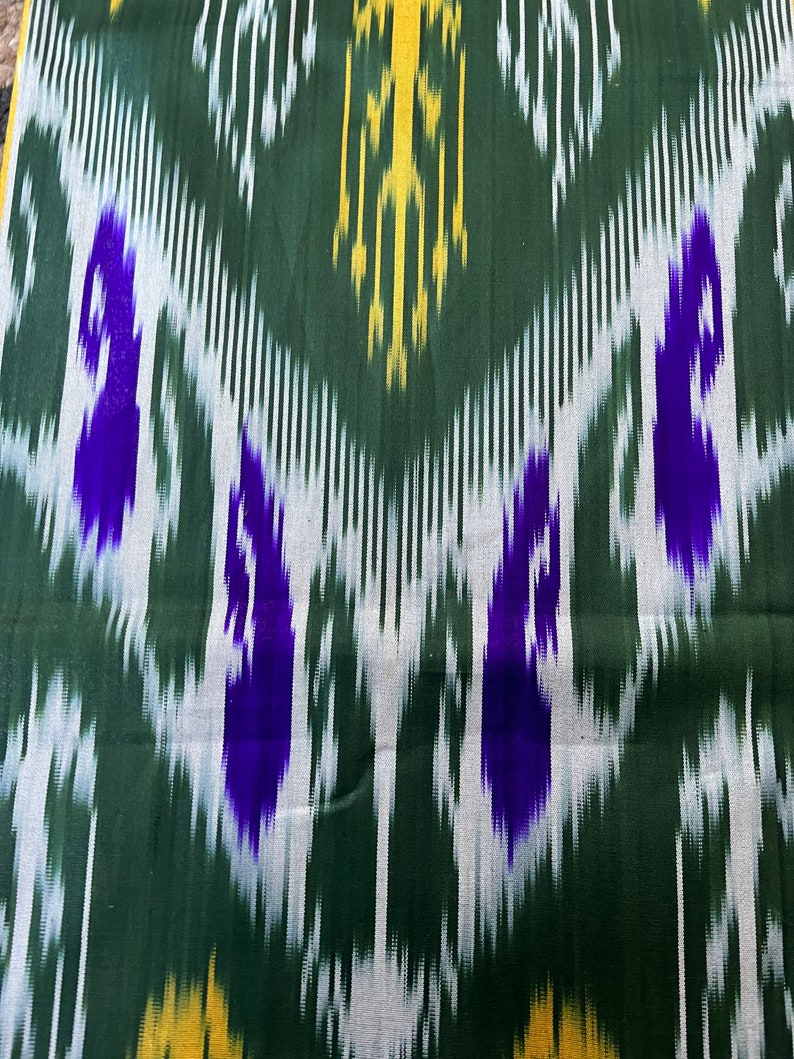 May include: Close-up of a fabric with a repeating ikat pattern. The design features a dark green background with vertical white and yellow lines. Bold purple shapes are interspersed throughout the pattern, creating a striking visual effect. The fabric has a textured appearance.