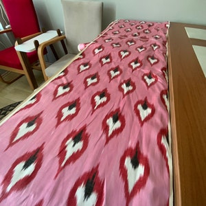 May include: Pink fabric with a white and red ikat pattern. The fabric is folded and laid on a wooden table.
