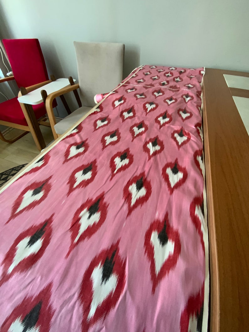 May include: Pink fabric with a white and red ikat pattern. The fabric is folded and laid on a wooden table.
