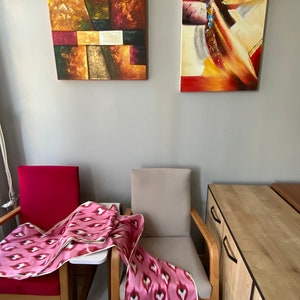 May include: Two abstract paintings hang on a wall above two chairs with pink and white patterned throws draped over them. A wooden cabinet with two drawers is in the background.