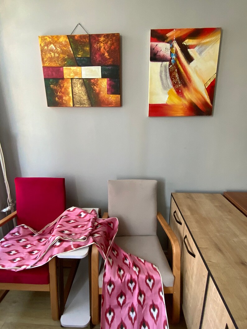 May include: Two abstract paintings hang on a wall above two chairs with pink and white patterned throws draped over them. A wooden cabinet with two drawers is in the background.