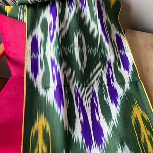 May include: A green fabric with a repeating pattern of white, purple, and yellow geometric shapes. The fabric is draped over a pink chair. The edges of the fabric are trimmed with a yellow border.
