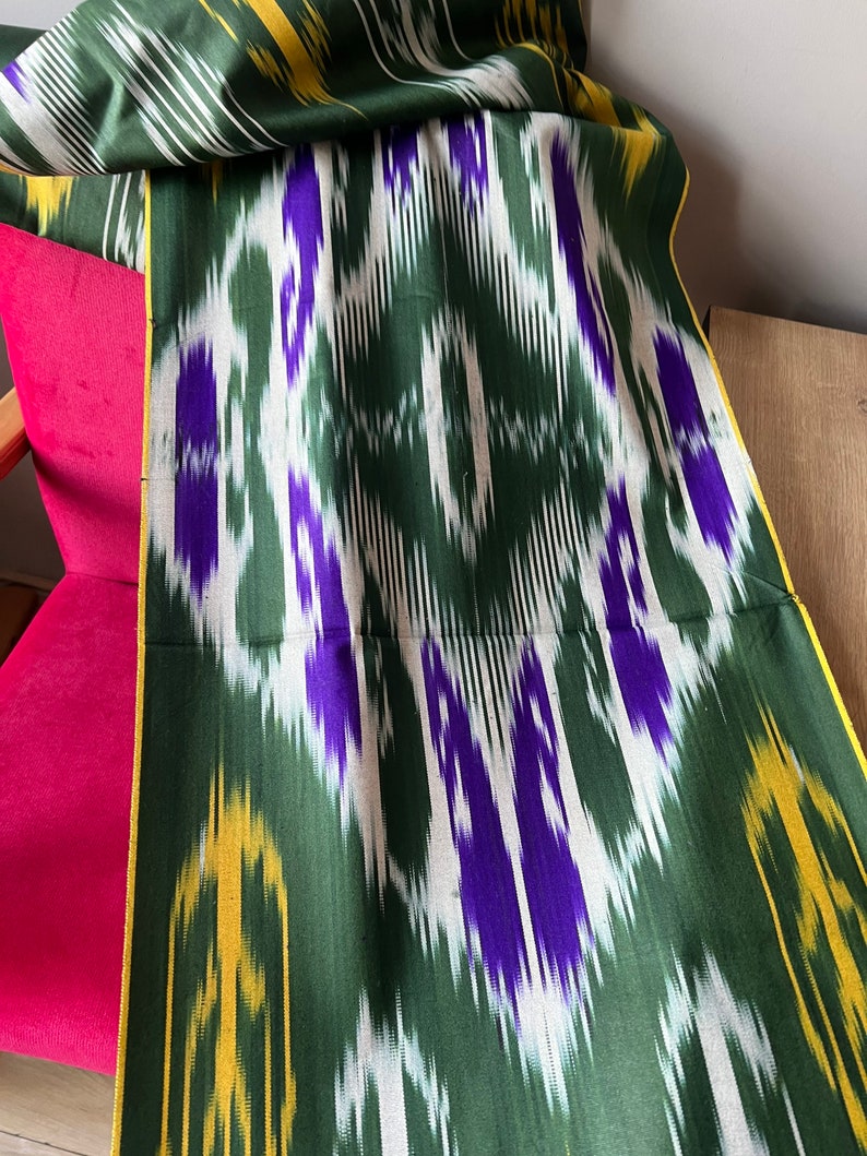 May include: A green fabric with a repeating pattern of white, purple, and yellow geometric shapes. The fabric is draped over a pink chair. The edges of the fabric are trimmed with a yellow border.