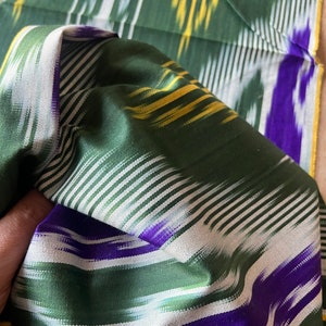 May include: Close-up of a folded green fabric with a striped pattern. The fabric features white, yellow, and purple stripes against a green background. The fabric appears to be silk or a similar material, with a slight sheen.
