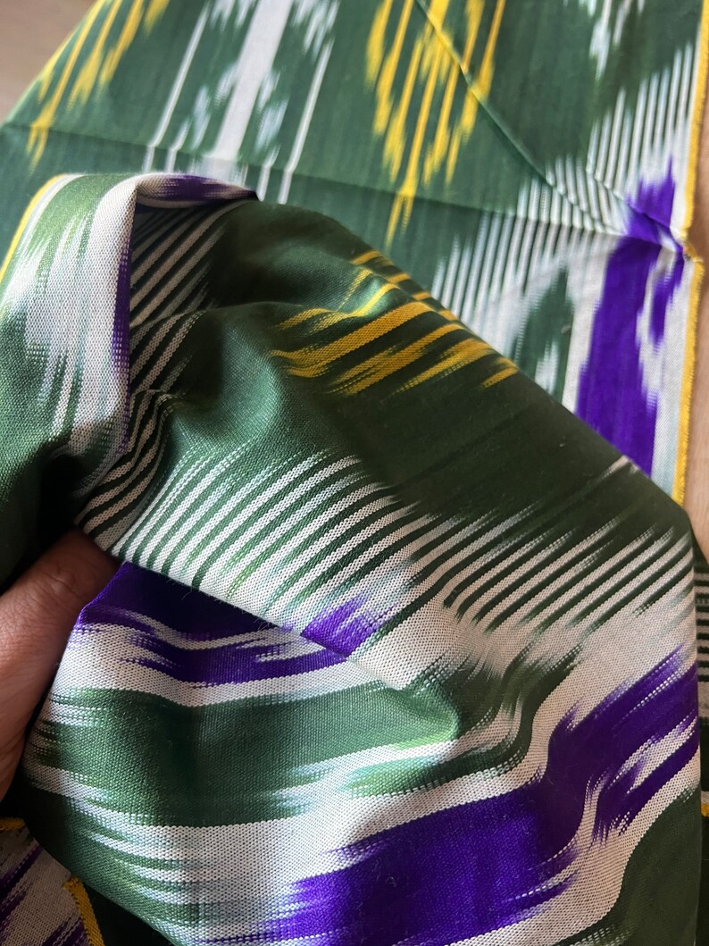 May include: Close-up of a folded green fabric with a striped pattern. The fabric features white, yellow, and purple stripes against a green background. The fabric appears to be silk or a similar material, with a slight sheen.