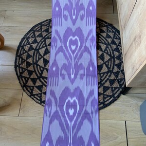 May include: A purple and white ikat fabric with a traditional pattern. The fabric is folded and lying on a woven rug.