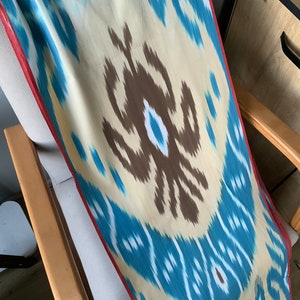 May include: A long, rectangular piece of fabric with a blue, white, and brown ikat pattern. The fabric has a red border and is draped over a wooden chair.