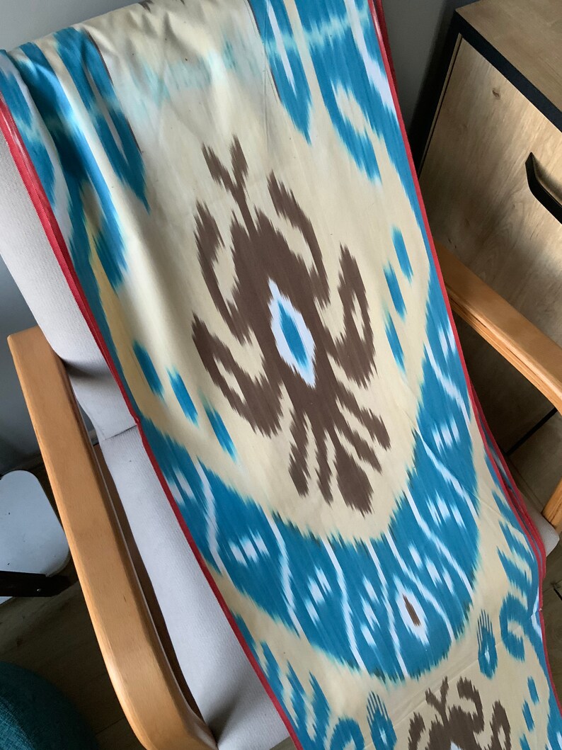 May include: A long, rectangular piece of fabric with a blue, white, and brown ikat pattern. The fabric has a red border and is draped over a wooden chair.