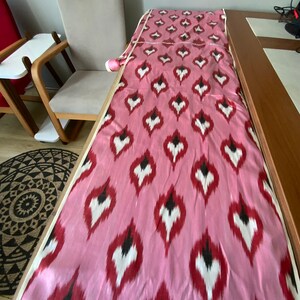 May include: Pink fabric with a white and black ikat pattern. The fabric is folded and laid out on a table.