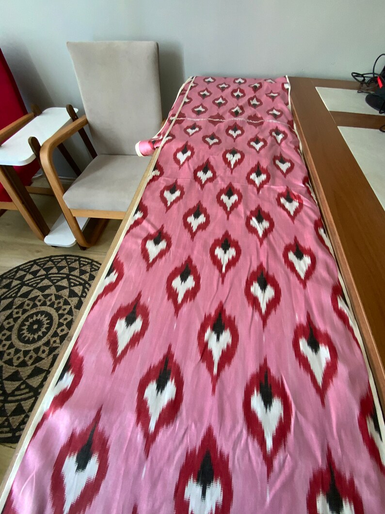 May include: Pink fabric with a white and black ikat pattern. The fabric is folded and laid out on a table.