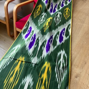 May include: A green textile with a repeating pattern of white, yellow, and purple shapes. The fabric has a yellow border and is draped over a wooden surface. The pattern has a geometric design.