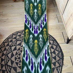 May include: A length of green fabric with a repeating pattern of white, yellow, and purple shapes. The fabric has a yellow border and is displayed on a wooden surface with a round, patterned rug.