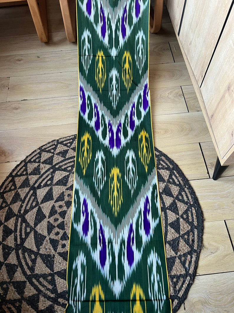 May include: A length of green fabric with a repeating pattern of white, yellow, and purple shapes. The fabric has a yellow border and is displayed on a wooden surface with a round, patterned rug.