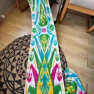 May include: A white fabric with a colourful ikat pattern. The pattern features green, blue, yellow, and pink colours. The fabric is folded in half and laid out on a wooden floor.