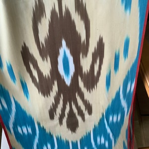 May include: A close-up of a fabric with a brown and blue ikat pattern. The fabric has a white background with brown and blue swirls and a red border.