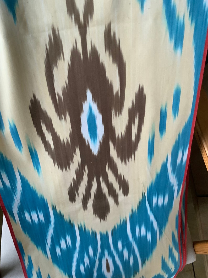 May include: A close-up of a fabric with a brown and blue ikat pattern. The fabric has a white background with brown and blue swirls and a red border.
