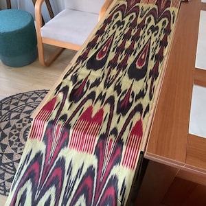 May include: A long, patterned textile with a repeating design of black, burgundy, and cream-colored shapes. The fabric is draped over a wooden surface, showcasing its intricate pattern and texture. The textile appears to be a table runner or decorative cloth.