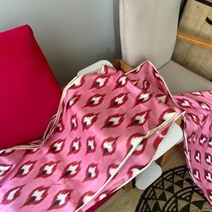 May include: Pink and white patterned fabric with a cream border. The fabric is folded and draped over a chair.