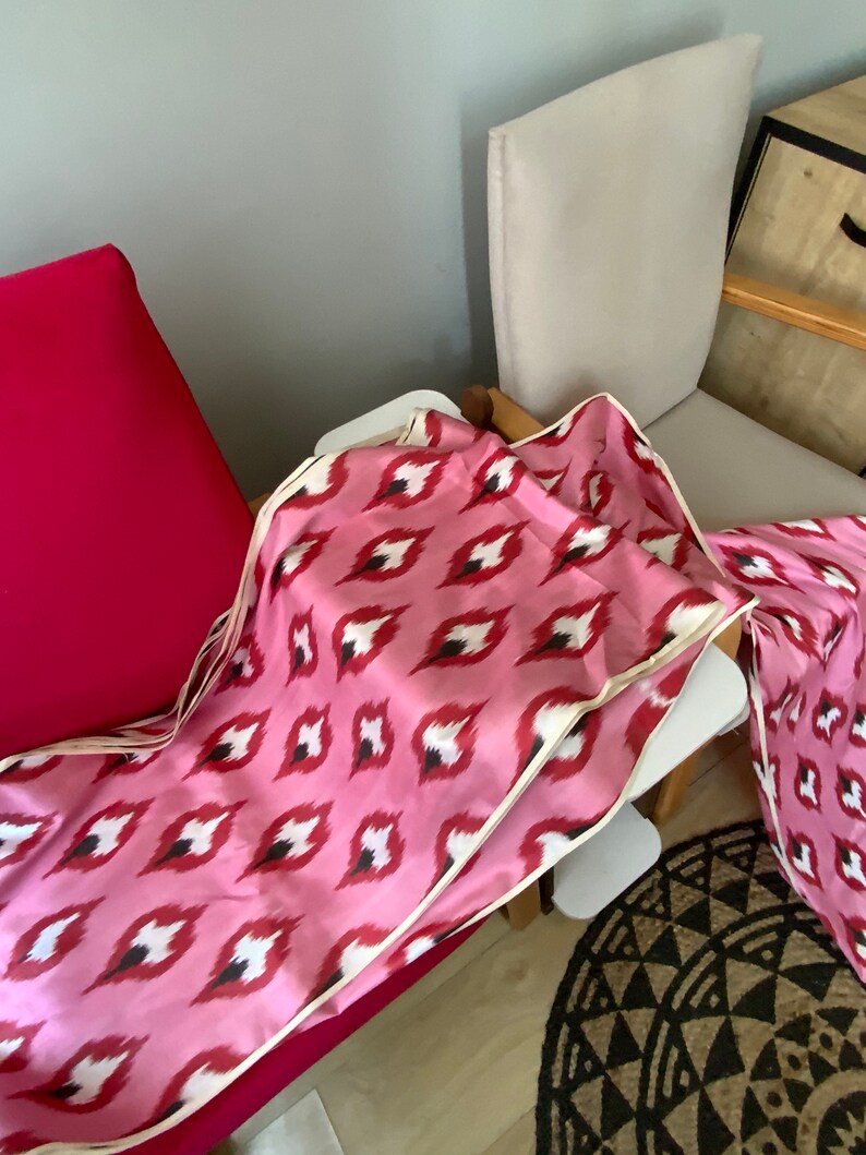 May include: Pink and white patterned fabric with a cream border. The fabric is folded and draped over a chair.
