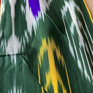 May include: Close-up of a green fabric with a vibrant ikat pattern. The design features vertical stripes in white, yellow, and purple. The fabric has a pocket detail with a green thread seam.