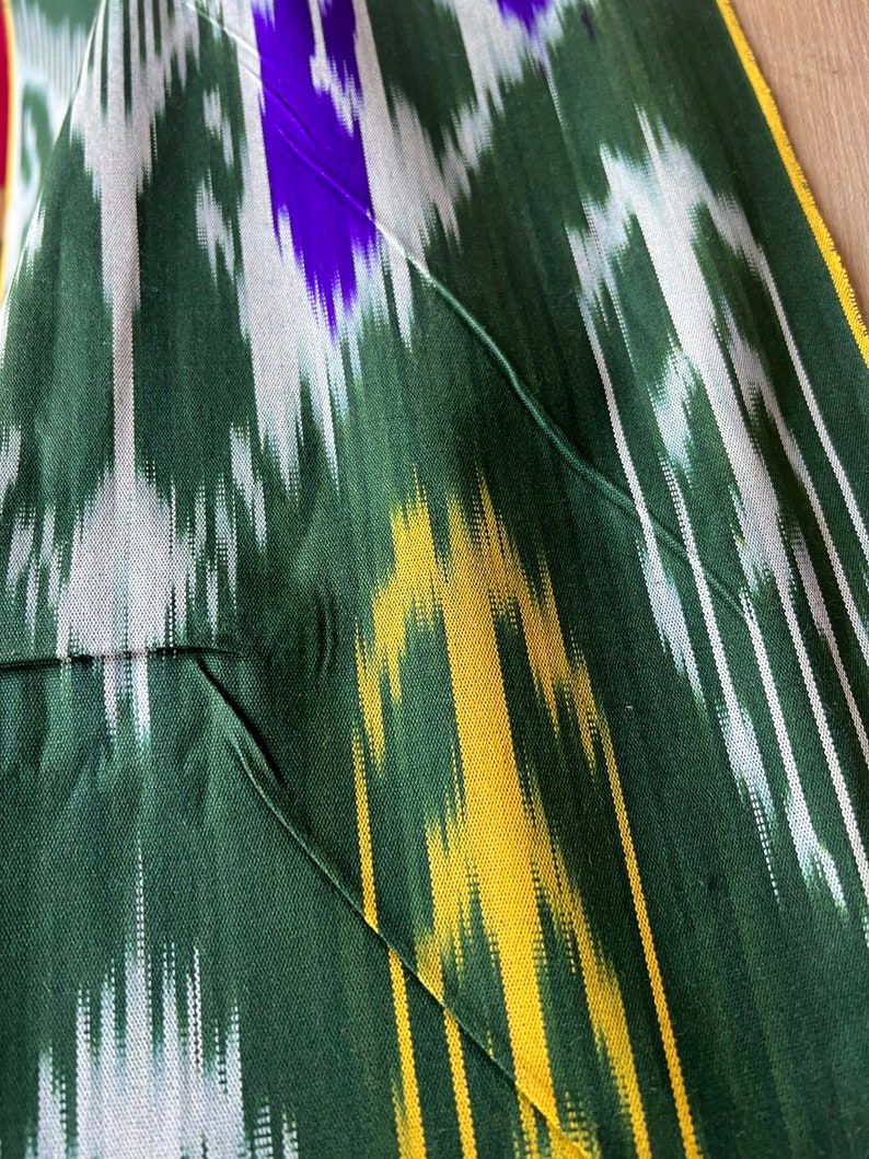 May include: Close-up of a green fabric with a vibrant ikat pattern. The design features vertical stripes in white, yellow, and purple. The fabric has a pocket detail with a green thread seam.
