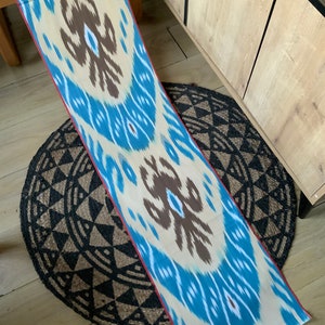 May include: A long, cream-colored fabric with a blue and brown ikat pattern. The fabric has a red border and is lying on a black and brown circular rug.