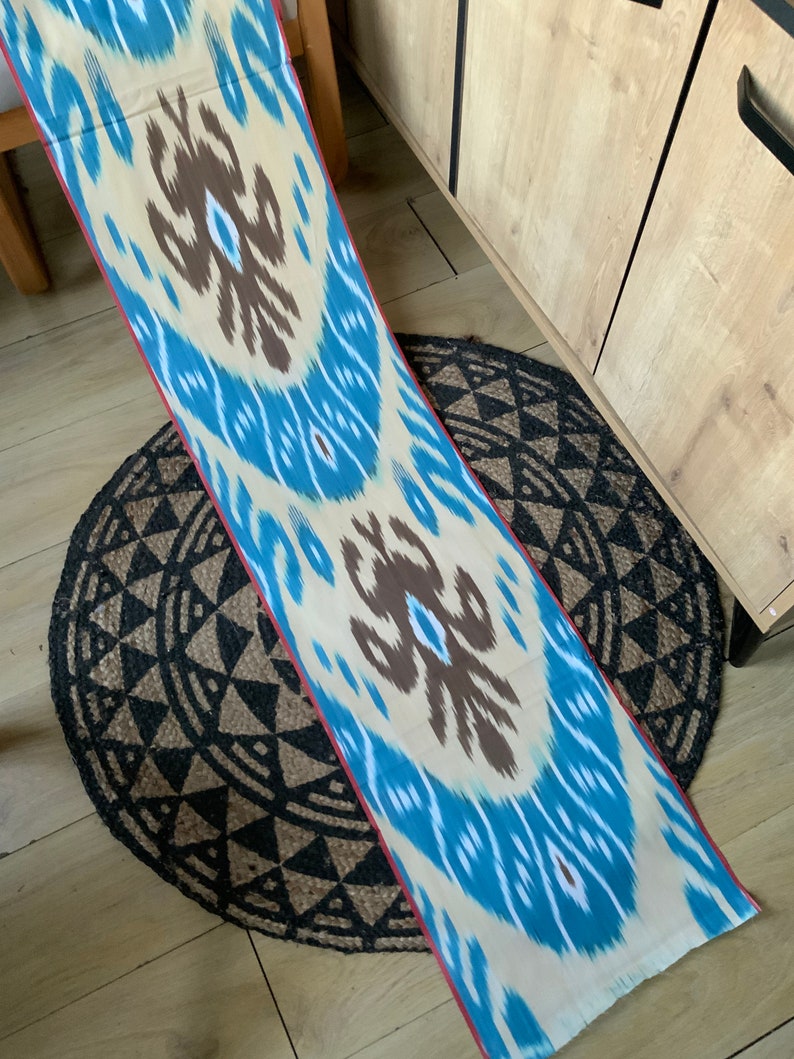 May include: A long, cream-colored fabric with a blue and brown ikat pattern. The fabric has a red border and is lying on a black and brown circular rug.