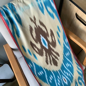 May include: A blue, white, and brown ikat fabric with a red border. The fabric is draped over a wooden chair.