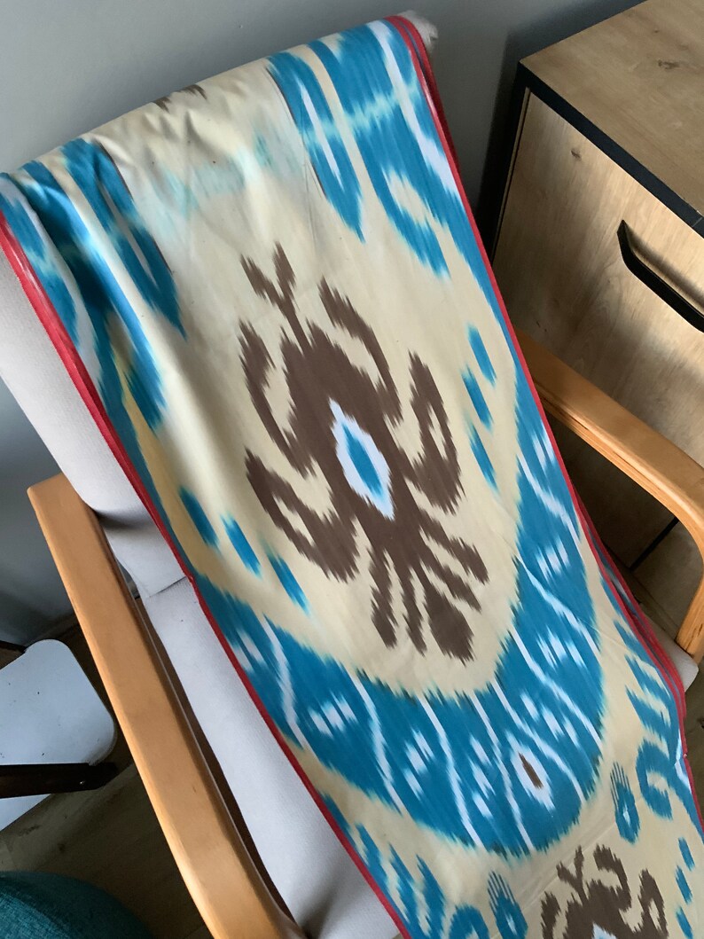 May include: A blue, white, and brown ikat fabric with a red border. The fabric is draped over a wooden chair.