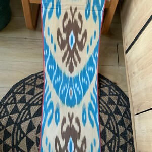 May include: A long, narrow piece of fabric with a blue, brown, and white ikat pattern. The fabric has a red border and is folded in half.