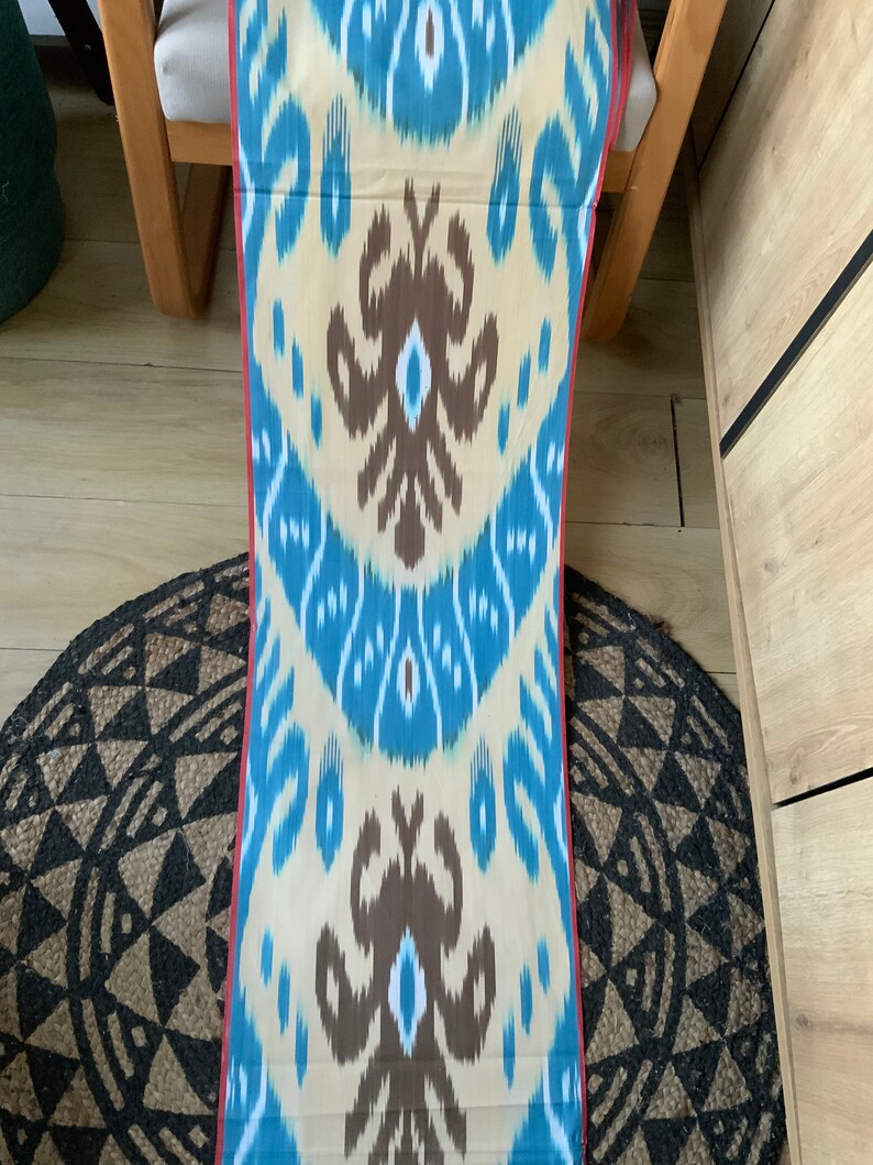 May include: A long, narrow piece of fabric with a blue, brown, and white ikat pattern. The fabric has a red border and is folded in half.