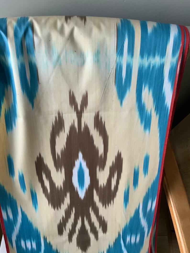 May include: A close-up of a fabric with a brown, blue, and white ikat pattern. The fabric has a red border.